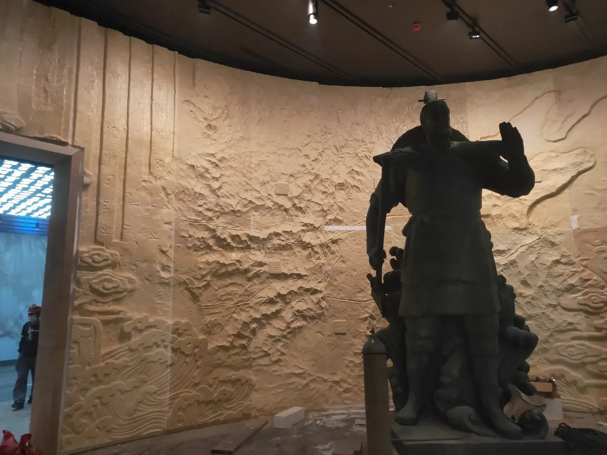 Bronze figure displayed in a gallery against a relief wall